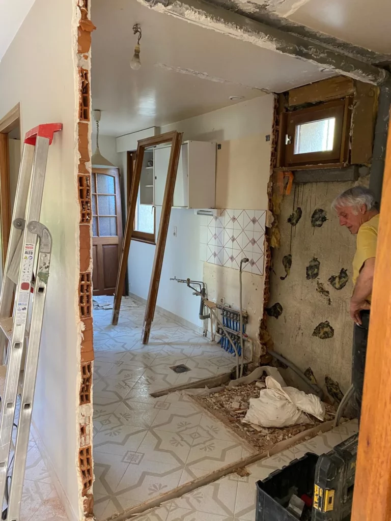 Vue intérieure d'un chantier de démolition d'un rez de chaussé à Gaillard. Le faux plafond a été retiré, laissant apparaître la structure supérieure en bois et les câbles électriques qui pendent. Le mur de séparation a été partiellement abattu, dévoilant un sol carrelé recouvert de poussière et de petits gravats.