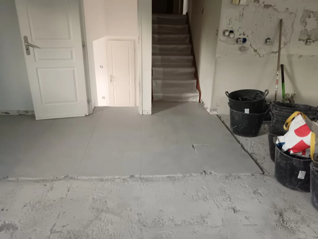 Démolition du carrelage d'une maison à Excenevex : salon vidé avec chape nue, seaux et porte vers escalier