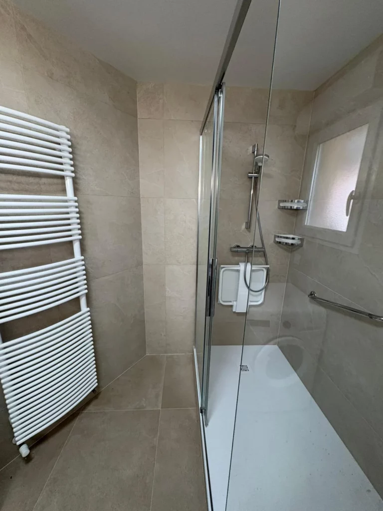 Détail de la douche rénovée à Annemasse avec carrelage beige, sèche-serviettes blanc et paroi vitrée chromée