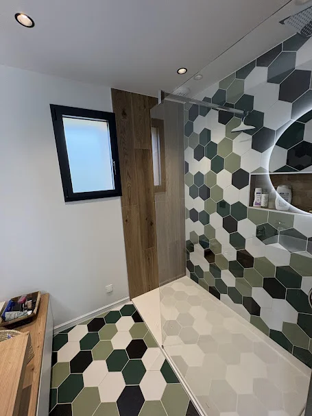 Vue d'une salle de bain avec vasque à poser blanche sur plan bois, miroir rond rétroéclairé et sol en carrelage hexagonal aux tons vert, kaki et blanc. Photographie illustrant une pose de carrelage dans une maison à Ballaison, avec un motif hexagonal graphique au sol et sur la paroi de douche.